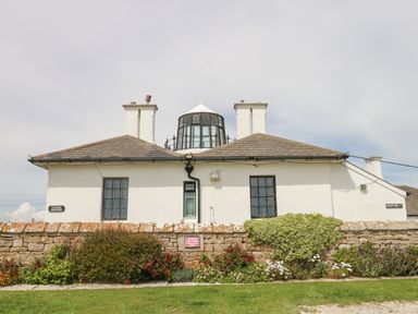 A white building with multiple windows and chimneys behind a stone wall with flowers at Old Higher Lighthouse Stopes Cottage in Portland Bill