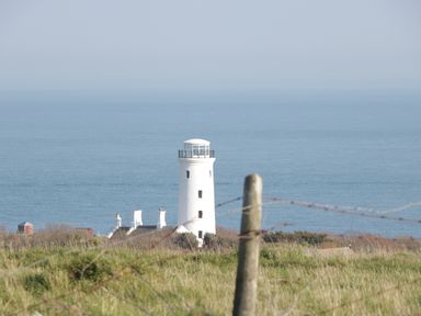 A white lighthouse and buildings near a grassy field with a post and barbed wire fence in the foreground at Old Higher Lighthouse Branscombe Lodge Portland Bill