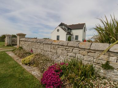 A white house behind a stone wall with flowerbeds and a grassy area at Old Higher Lighthouse Branscombe Lodge Portland Bill