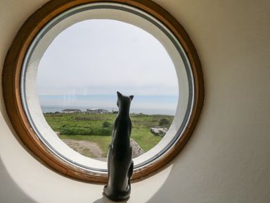 A round window with a black cat statue looking out at green fields and buildings at Old Higher Lighthouse Branscombe Lodge in Portland Bill