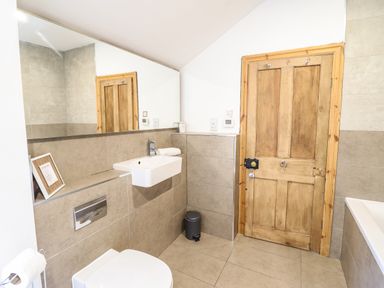 A bathroom with a wooden door, a wall-mounted sink, a toilet, a large mirror, and a small trash bin at 3 Railway Cottages in Betws-y-Coed