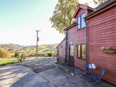 A red wooden house with two chairs and a small table outside overlooking a green hill landscape at Cedar Cottage in Llanidloes
