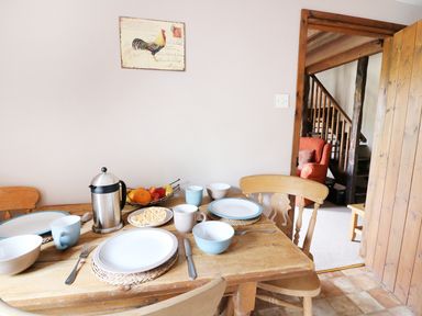 A dining table set with plates bowls mugs and fruit bowl with a roosting chicken picture on the wall at Beech Cottage in Llanidloes