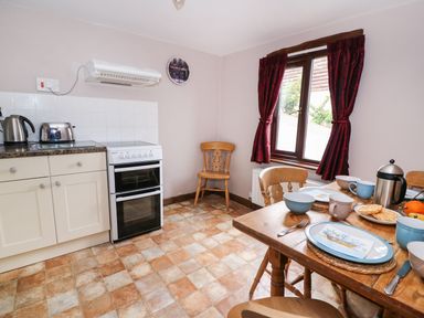 A kitchen with a wooden dining table set with dishes and utensils near a window with red curtains and a stove next to a countertop with a kettle and toaster at Beech Cottage in Llanidloes
