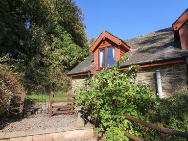 Exterior of a stone house with a slate roof and wooden windows surrounded by greenery and a wooden gate at Oak Cottage in Llanidloes