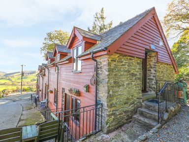 A row of houses with red wooden siding and stone walls on a hillside with outdoor seating at Oak Cottage in Llanidloes