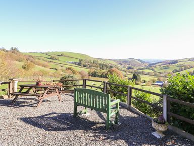 An outdoor seating area with a green bench and wooden picnic table overlooking green hills and farmland at Oak Cottage in Llanidloes