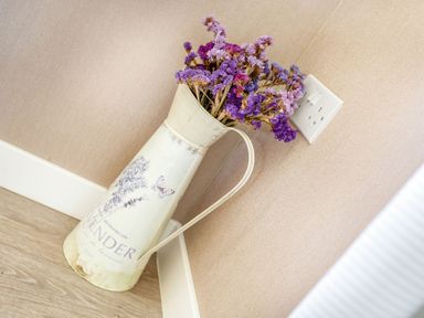 A metal pitcher with dried purple flowers on the floor against a beige wall with a white electrical outlet at Lavender Lodge in Charlton Horethorne near Sherborne
