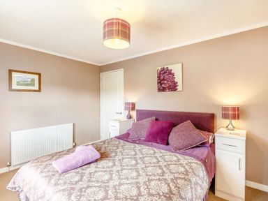A bedroom with a double bed with purple bedding and pillows two white bedside cabinets with lamps a framed picture and a radiator at Lavender Lodge in Charlton Horethorne near Sherborne