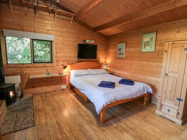 A bedroom with a wooden bed with blue towels on it a wooden wardrobe a window a wall-mounted television and a wood-burning stove at The Log Cabin in Oban