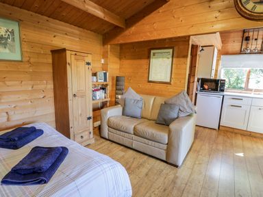 A room with a bed with towels a beige sofa a wooden wardrobe and a kitchen area at The Log Cabin in Oban