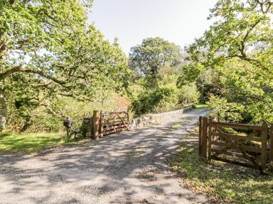 A gravel driveway with wooden gates and a stone wall surrounded by trees at The Log Cabin in Oban