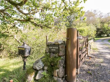 A gravel driveway with a wooden gate and a stone pillar with a Glen EuChar House sign next to a mailbox surrounded by trees at The Log Cabin in Oban