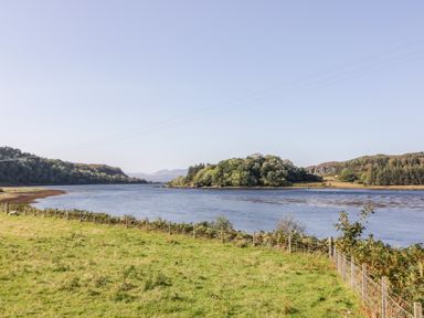 A river with green hills and trees on both sides and a grassy field with a fence in the foreground at The Log Cabin in Oban