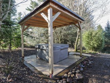 A wooden gazebo with a covered hot tub in a garden surrounded by trees at The Log Cabin in Oban