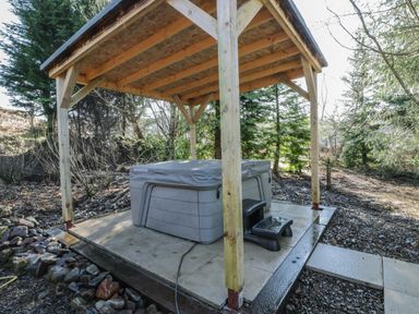 An outdoor hot tub with cover under a wooden shelter in a wooded area at The Log Cabin in Oban