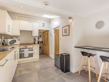 A kitchen with white cabinets an oven a microwave two trash bins and a breakfast bar with two stools at 2 Bryn Tirion in Llan Ffestiniog