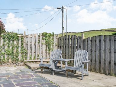 Two wooden chairs with a small table between them on a stone patio with a wooden fence and green hills in the background at 2 Bryn Tirion in Llan Ffestiniog
