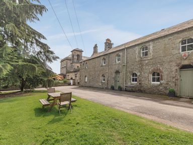 A stone building with multiple windows next to a grassy area with a wooden picnic table and chairs at Reiver's Rest in Hesleyside near Bellingham