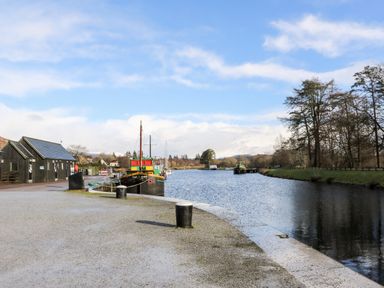 A riverside with boats docked near black buildings and trees along the banks at Glen Dessary in Corpach near Fort William
