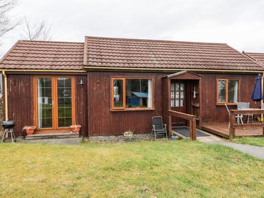 A wooden house with a tiled roof and glass door next to windows and outdoor chairs on grass at Glen Dessary in Corpach near Fort William