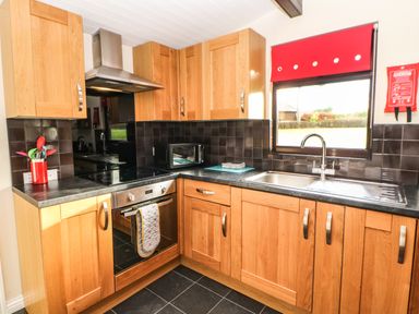 A kitchen with wooden cabinets, a stove with an oven, a stainless steel sink under a window with a red blind, and black tiled backsplash at Primrose Lodge in Saltburn-by-the-Sea