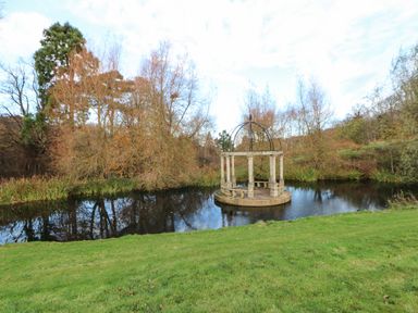 A small pond with a circular stone gazebo in the center surrounded by trees and grass at Primrose Lodge in Saltburn-by-the-Sea