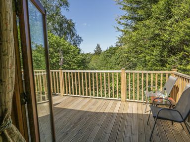 A wooden deck with two chairs and a small table with a bottle and glasses surrounded by trees at Striding Edge White Cross Bay near Troutbeck Bridge