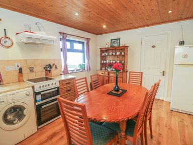 A kitchen with a wooden dining table and chairs a washing machine an oven a window with checkered curtains and a wooden cupboard with dishes at Nanas House in Allihies County Cork