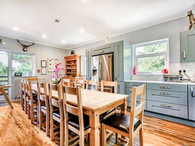 A kitchen with a long wooden dining table surrounded by chairs and gray cabinets with a stainless steel refrigerator at Eden House in Bardsea
