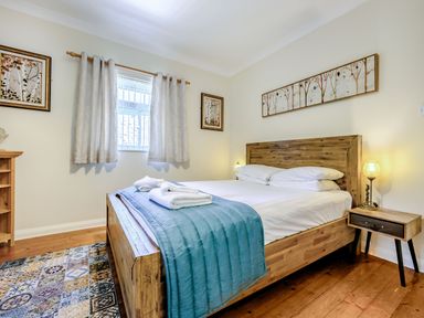 A bedroom with a wooden bed with white linens and teal blanket next to two framed pictures a window with curtains and a small wooden nightstand with a lamp at Eden House in Bardsea