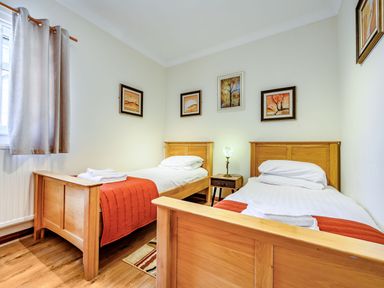 A bedroom with two wooden single beds with white bedding and orange throws a small wooden nightstand with a lamp and framed paintings on the walls at Eden House in Bardsea