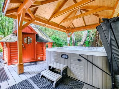 An outdoor hot tub under a wooden gazebo next to a small orange wooden cabin surrounded by greenery at Eden House in Bardsea
