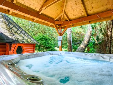 A bubbling hot tub under a wooden gazebo with trees and a small wooden cabin nearby at Eden House in Bardsea