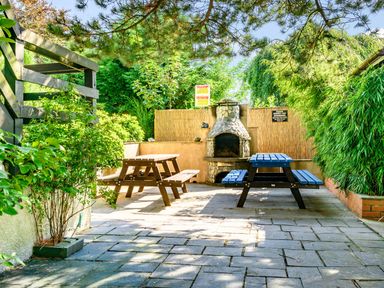 An outdoor patio with two wooden picnic tables and a stone barbecue grill surrounded by green plants at Eden House in Bardsea