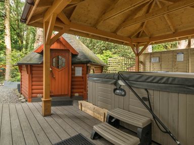 An outdoor wooden hot tub with steps under a wooden pergola and a small red wooden cabin in a forested area at Eden House in Bardsea