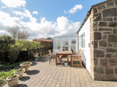 An outdoor patio area with a wooden table and chairs next to a house with a stone wall and glass doors at Lyndhurst Cottage in Beadnell