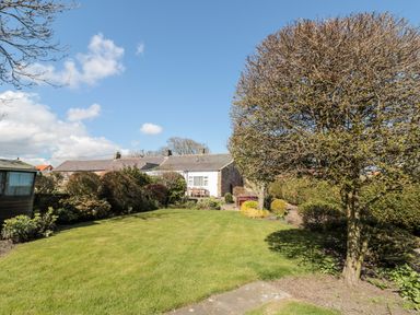 A garden with a trimmed lawn trees shrubs and a stone building in the background at Lyndhurst Cottage in Beadnell