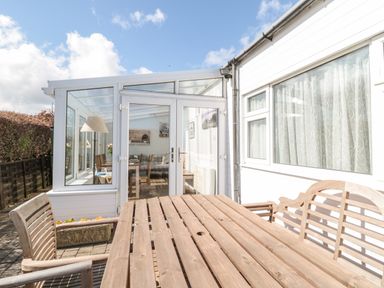 An outdoor wooden table and chairs on a patio next to a white conservatory with glass doors and windows at Lyndhurst Cottage in Beadnell