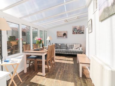 A sunroom with a wooden dining table and chairs a plaid sofa wooden bench and floor lamp at Lyndhurst Cottage in Beadnell