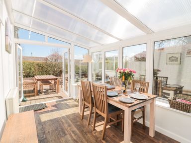 A dining area with a wooden table and chairs set for four inside a sunroom opening to an outdoor patio with wooden furniture at Lyndhurst Cottage in Beadnell