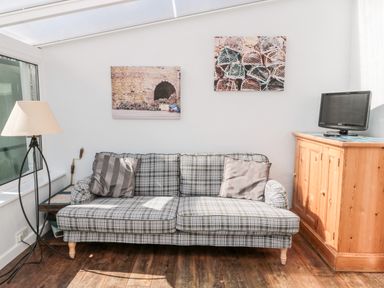 A plaid sofa with two cushions next to a wooden cabinet with a small TV on top and a floor lamp in a bright room at Lyndhurst Cottage in Beadnell