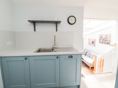 A kitchen sink with blue cabinets a wall clock and a shelf above and a view into a living area with a couch and artwork at Lyndhurst Cottage in Beadnell