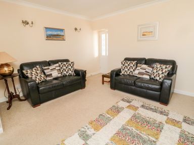 A living room with two black leather sofas with cushions a small wooden side table with a lamp a patterned rug and framed pictures on the walls at Lyndhurst Cottage in Beadnell