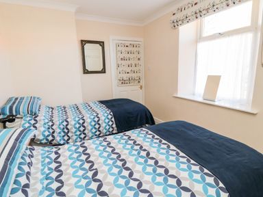 A bedroom with two single beds with patterned bedding a mirror on the wall and a window with a small cloth on the sill at Lyndhurst Cottage Beadnell