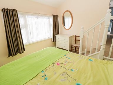 A bedroom with a bed covered in a green floral bedspread a white dresser a wooden chair a round mirror and a white staircase at Lyndhurst Cottage in Beadnell
