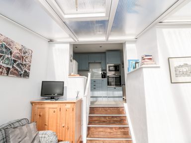 A small living area with a wooden cabinet holding a TV next to stairs leading to a kitchen with blue cabinets at Lyndhurst Cottage in Beadnell
