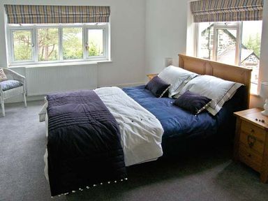 A bedroom with a bed with dark blue and white bedding two windows with striped curtains a white chair and wooden bedside tables at Llechwedd in Dolgellau