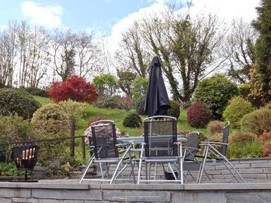 An outdoor patio with metal chairs and a table with a closed umbrella surrounded by shrubs and trees at Llechwedd in Dolgellau