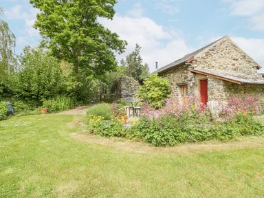A stone cottage with a red door surrounded by grass trees and a flower garden at The Shippon in Llechwedd The Conwy Valley